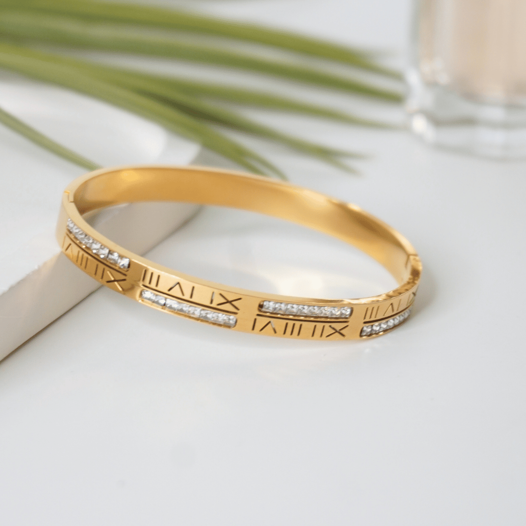 Gold bracelet with Roman numeral design on a light surface with green leaves in the background